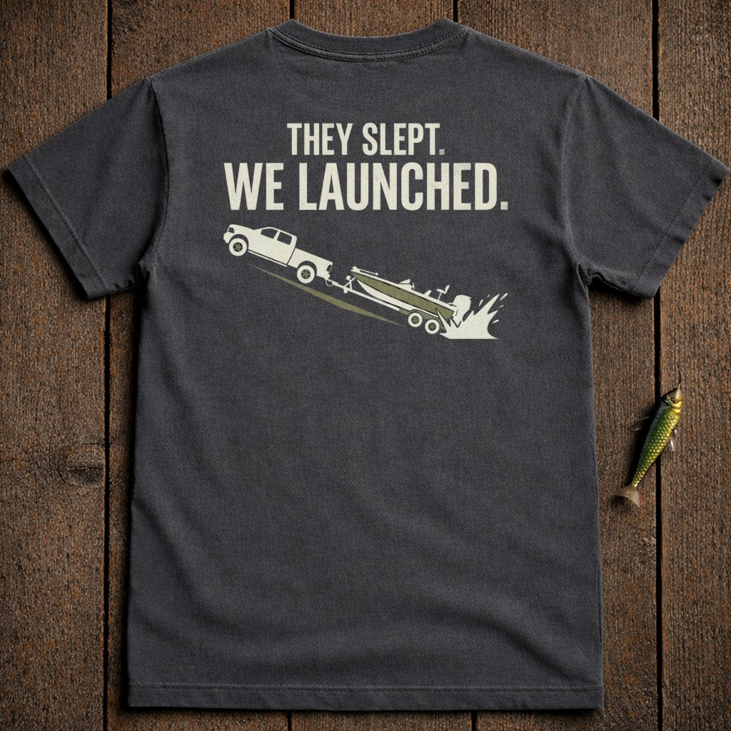 Dark t-shirt with white graphic of truck launching boat, text "THEY SLEPT. WE LAUNCHED."