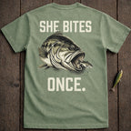 Green t-shirt with large bass fish biting smaller fish, text "SHE BITES ONCE."