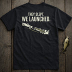 Black t-shirt graphic: truck launching boat into water with text "THEY SLEPT. WE LAUNCHED."