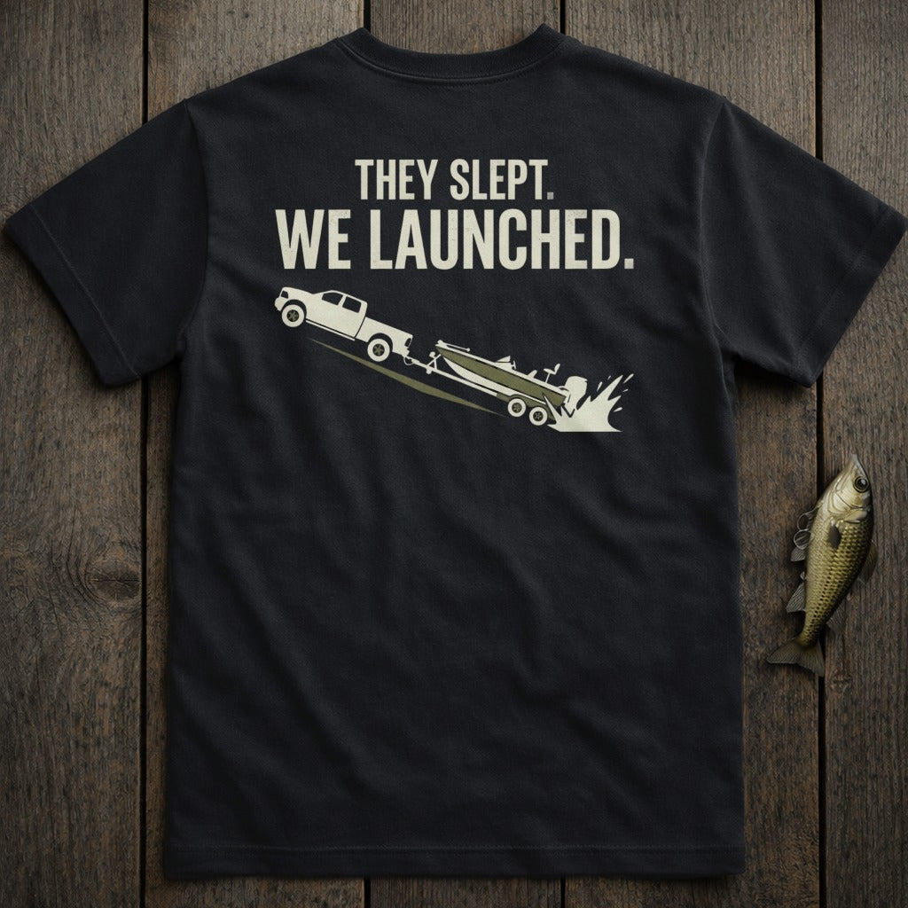 Black t-shirt graphic: truck launching boat into water with text "THEY SLEPT. WE LAUNCHED."